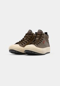 Brown high-top sneakers with a textured finish, cream rubber toe cap, black rubber sole, and contrasting red logo tag on the side.