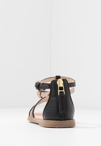 Svart lädersandal med guld zipperedetalj bak, med flera tunna remmar och en beige accentrem. Platt sula.
