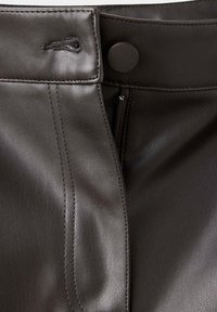 Gros plan sur la taille d’un pantalon en cuir marron foncé montrant un bouton, une fermeture éclair et des coutures détaillées sur la ceinture.