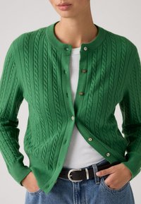 Femme portant un cardigan boutonné en maille torsadée verte par-dessus un haut blanc côtelé, associé à un jean bleu et une ceinture noire avec une boucle argentée.