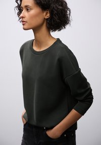 Ciemnozielony sweatshirt z okrągłym dekoltem, długimi rękawami i luźnym krojem. Gładka tekstura, minimalistyczny design, noszony z czarnymi spodniami.