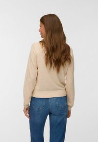 Frau mit langen braunen Haaren, die einen hellbeigen Pullover und blaue Jeans trägt, steht mit dem Rücken zu einem schlichten Hintergrund.