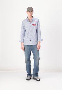 Striped button-up shirt with red logo patch, denim jeans, and gray cap. Shoes feature a sporty design in silver and black accents.