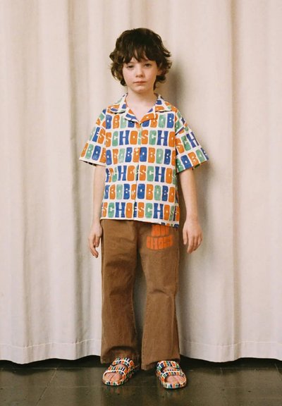 Colorful short-sleeve button-up shirt featuring large block letters in orange, blue, green; brown wide-leg pants with printed accents; multicolored sandals.