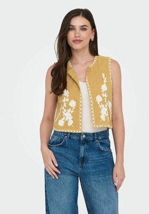 Senfgelbes Cropped-Top mit weißer Blumenstickerei, ärmelloses Design und gewelltem Saum, kombiniert mit hoch taillierten blauen Jeans.