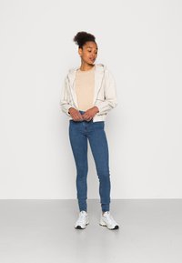 Jaqueta com fecho éclair de tom bege claro, com capuz, sobre uma blusa listrada de tom castanho claro, combinada com calças jeans skinny azuis e ténis brancos.