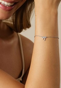 Bracelet en argent présentant un délicat design de nœud avec de petits cristaux, porté sur un poignet bronzé, mettant en valeur le métal brillant et les accents détaillés.
