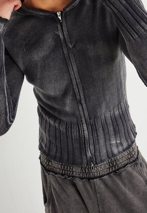 Zbliżenie na osobę ubrany w ciemnoszary sweter na zamek wykonany z dzianiny z prążkowanym dołem oraz dopasowane teksturowane spodnie.