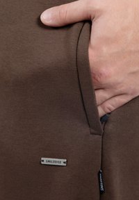 Indumento marrone in cotone con una tasca laterale. È visibile una targhetta d'argento con la scritta "SMILODOX". Texture liscia e design aderente sono caratteristiche notevoli.