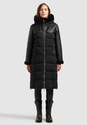 Femme portant un long manteau matelassé noir avec manches en cuir, capuche et poignets bordés de fourrure, pantalon noir et bottes noires, debout sur fond gris.