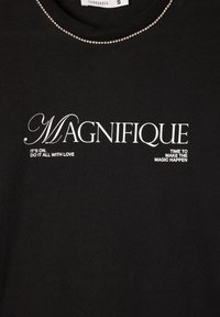 Czarna koszulka z bawełny z okrągłym dekoltem, z białym napisem i akcentem z cyrkoniami wzdłuż kołnierza. Napis zawiera "MAGNIFIQUE."