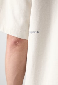 Krämfärgad bomull T-shirt med rullade ärmar, har liten text "scispiritual" i grått på vänster sida. Slen textur, avslappnad design.
