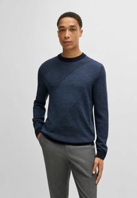 Pullover in maglia blu navy con un motivo diagonale testurizzato, scollatura a girocollo e polsini a costine, abbinato a pantaloni eleganti grigi chiari.