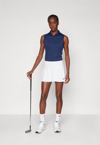 Námorně modré bezrukávové polo tričko s knoflíky, bílé skladané sukně, bílé golfové boty a ponožky s černým logem, držící golfovou hůl.