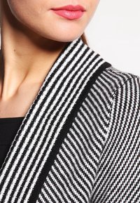 Gestreifter Pullover in Schwarz und Weiß mit einem Schalkragen, der ein strukturiertes Strickmuster aufweist. Der Kragen hat einen einfarbigen schwarzen Rand.