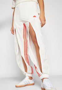 Henkilö, jolla on päällä valkoiset Adidas-urheiluhousut, joissa on punaiset raidat ja sivuvetoketjun avaukset, yhdistettynä valkoisiin lenkkareihin, joissa on kumipohjat.