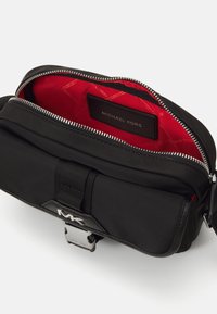 Borsa nera in nylon con interno rosso, chiusura a zip e accessori in argento. Presenta una tasca frontale e un logo impresso sul davanti.