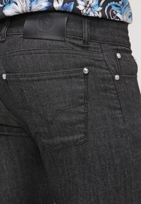 Jean en denim noir avec rivets métalliques, poche arrière et écusson de marque en cuir noir sous une chemise bleue à fleurs blanches.