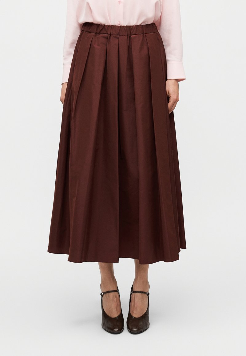 Καφέ, πλισέ μίντι φούστα από λείο ύφασμα, με ελαστική μέση. Συνδυάζεται με σκουρόκαφε παπούτσια.