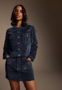 Ensemble deux pièces en velours bleu marine foncé, comprenant une veste boutonnée avec deux poches poitrine et une jupe assortie avec des poches latérales.