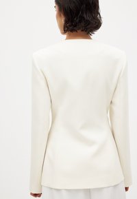 Balts pielāgots žakets ar garām piedurknēm, strukturētām plecu daļām un gludu audumu. Iezīmējas ar tīru dizainu un minimāliem šuvēm aizmugurē.