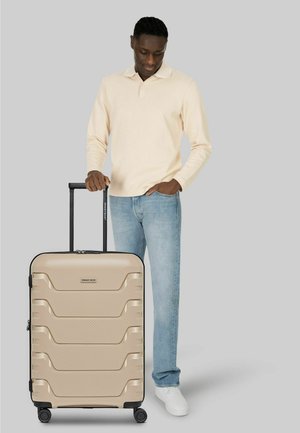 Uomo con camicia beige a maniche lunghe e jeans che tiene la maniglia di una valigia rigida beige con quattro ruote su sfondo grigio.