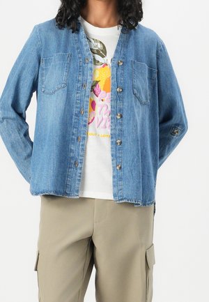 Person iført en knappet op blå denimskjorte over en hvid t-shirt med frugtmotiv og beige cargobukser.