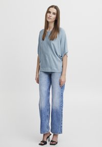 Lyseblå oversized top med korte ærmer, parret med falmede blå højtaljede bukser med bell-bottoms, suppleret af sorte hæle-sandaler.