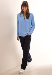 Camicia azzurro chiaro a maniche lunghe con scollatura henley, abbinata a jeans scuri e sneakers bianche, in piedi davanti a uno sfondo neutro.