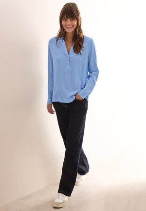 Lys blå langærmet skjorte med henley-udskæring, parret med mørke jeans og hvide sneakers, stående mod en neutral baggrund.