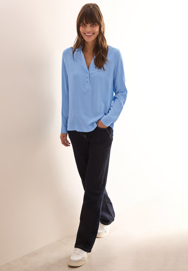 Camicia azzurro chiaro a maniche lunghe con scollatura henley, abbinata a jeans scuri e sneakers bianche, in piedi davanti a uno sfondo neutro.