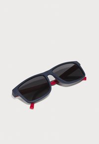 Ochelari de soare cu un cadru din plastic mat de culoare albastră închis și vârfuri roșii ale templelor. Lentile întunecate, formă unghiulară, fără logo-uri sau modele vizibile.