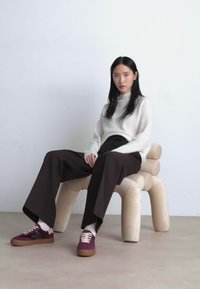 Bekväm sittdesign med en rundad, plysch, ljusfärgad stol. Modellen bär en vit ribbad tröja, bruna vida byxor och flerfärgade sneakers.