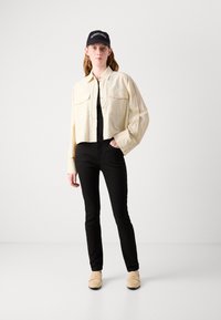 Crème cropped button-up shirt, zwarte top, zwarte skinny jeans en beige instappers, aangevuld met een zwarte pet met tekst.