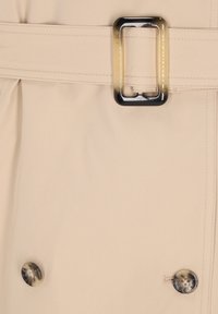 Beżowa tkanina płaszcza typu trench o gładkiej fakturze, z dużą czarno-kremową sprzączką paska oraz okrągłym brązowym guzikiem.