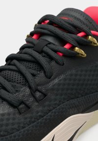 Sort atletisk sko med tekstureret mesh-overdel, pink detaljer og gyldne snørehuller. Har tykke sorte snørebånd og et markant buet således design.