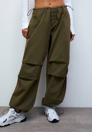 Persoon draagt losse olijfgroene cargobroek met trekkoorden en witte chunky sneakers, staand tegen een lichtgrijze muur.