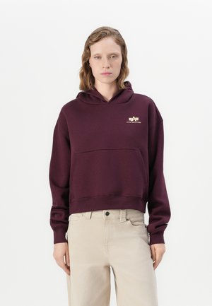 Kvinne iført en mørk lilla hettegenser med en liten gul logo og lysebeige bukser, stående mot en ensfarget hvit bakgrunn.
