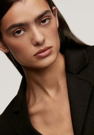 Joven con cabello largo y oscuro, vistiendo un abrigo negro y pequeños pendientes de oro, mirando directamente a la cámara con una expresión neutral.