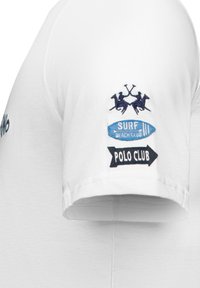 Biała bawełniana koszulka z krótkim rękawem. Posiada haftowane naszywki: dwóch graczy polo, niebieski napis "SURF BEACH CLUB" oraz czarny banner "POLO CLUB".
