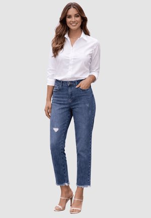 Mujer sonriente con cabello largo, vistiendo una camisa blanca abotonada, jeans azules rasgados con dobladillos deshilachados y sandalias de tacón beige.