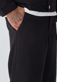 Pantalon noir avec une poche latérale, présentant une texture lisse et une coupe ajustée. L'ourlet d'une chemise blanche est visible à la taille.