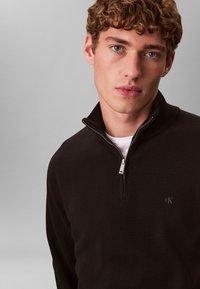 Joven con pelo rizado que lleva un suéter negro con cremallera hasta un cuarto sobre una camisa blanca, mirando directamente a la cámara sobre un fondo gris liso.