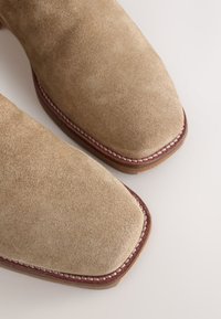 Khakifarbene Wildlederschuhe mit quadratischer Spitze, versehen mit einem kontrastierenden braunen Lederbesatz und sichtbaren, entlang der Sohle genähten Details.