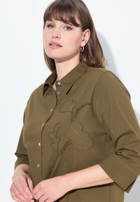 Blusa verde oliva con colletto a camicia, maniche 3/4 e ricami floreali in marrone e beige sul davanti. Texture morbida.