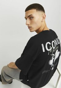 Czarna bawełniana koszulka z oversize'owym krojem. Zawiera białe napisy "ICONIC" na plecach oraz grafikę. Model siedzi na krześle, ubrany w szare dżinsy.