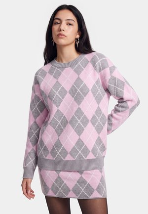Vrouw draagt een bijpassende roze en grijze argyle trui en rok met donkere panty's en creolen op een lichte achtergrond.