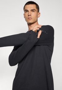 Chemise à manches longues noire en tissu côtelé avec des poignets à trou pour le pouce et un col rond, affichant une coupe décontractée et des détails de broderie subtils.