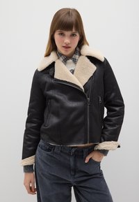 Giacca di shearling nera con colletto beige, chiusura frontale con zip e tasche laterali. Sono visibili anche pantaloni in velluto a coste grigi con finitura testurizzata.