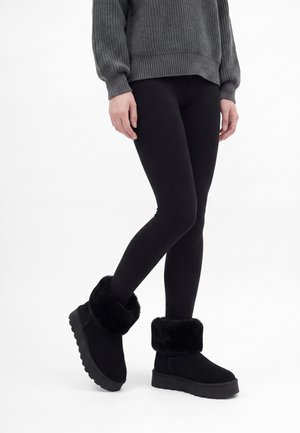 Personne portant des leggings noirs, un pull en maille gris et des bottes d'hiver noires doublées de fourrure, debout avec les jambes croisées.